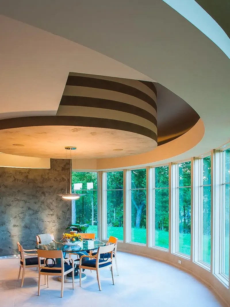 Round dinette under a striped drum ceiling with a curved wall of tall windows to the trees