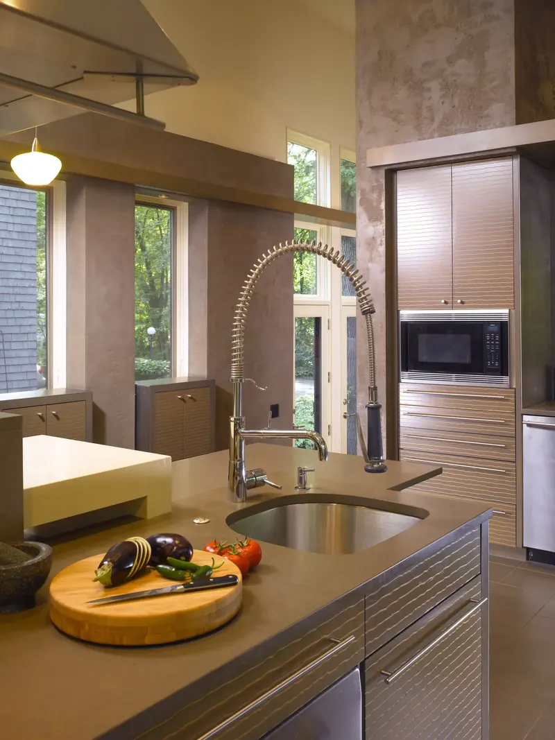 Island prep sink with spring faucet and cutting board, tall cabinets and windows to the trees beyond