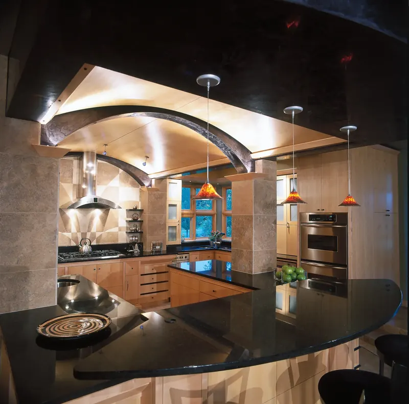 Curved black granite island under amber pendants and an arched beam, light wood cabinets beyond