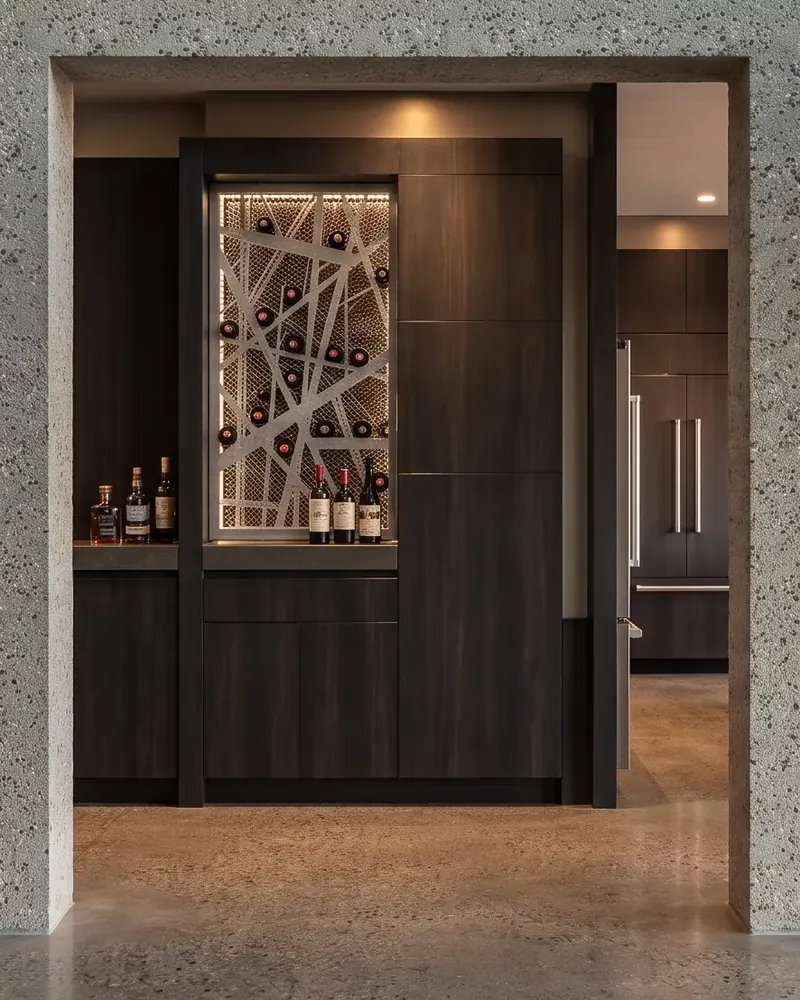 Dark wood cabinetry with a lit wine rack behind an abstract metal grille, framed by a concrete opening