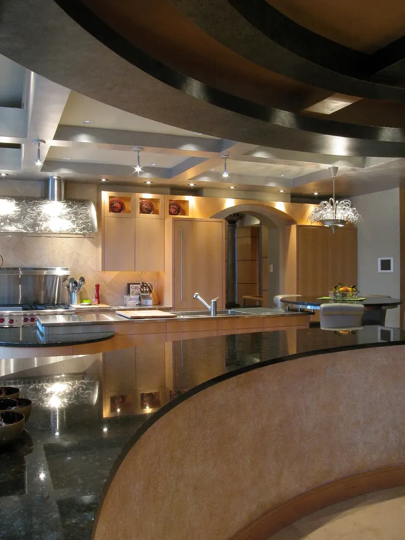Kitchen seen past a curved black stone counter, with layered ceilings and light wood cabinets