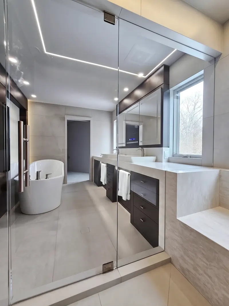 Pale stone bathroom seen through a glass shower, with a long stone-topped vanity and freestanding tub, Minneapolis