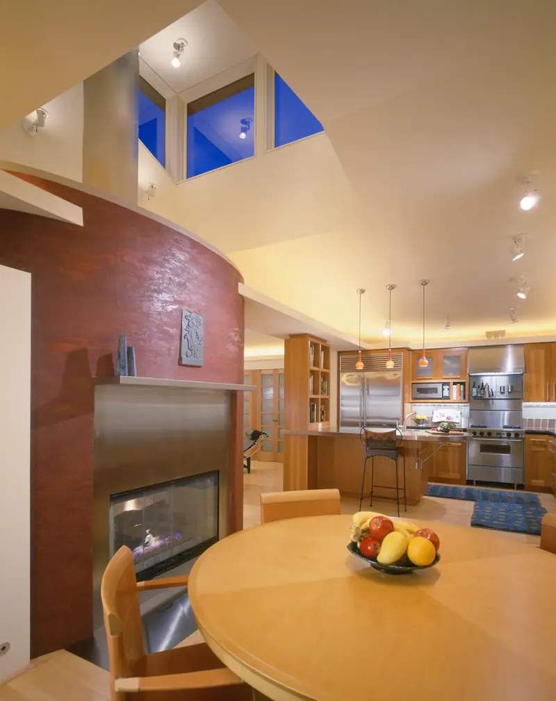 Curved red plaster wall with a stainless fireplace dividing a dining table from the kitchen