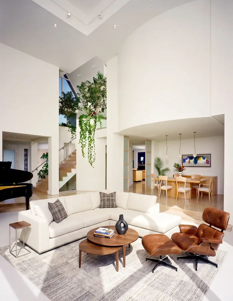 Double-height living room with grand piano, white sectional and leather lounge chair, open to the planted atrium and dining room