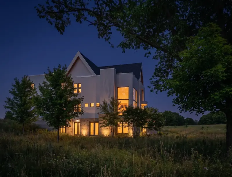 The Winter Green Atrium at dusk, warm light glowing from tall windows of the white gabled house beyond a dark meadow