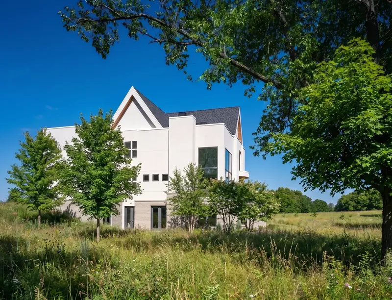 White gabled house in daylight seen across a meadow of tall grasses, framed by an overhanging tree branch